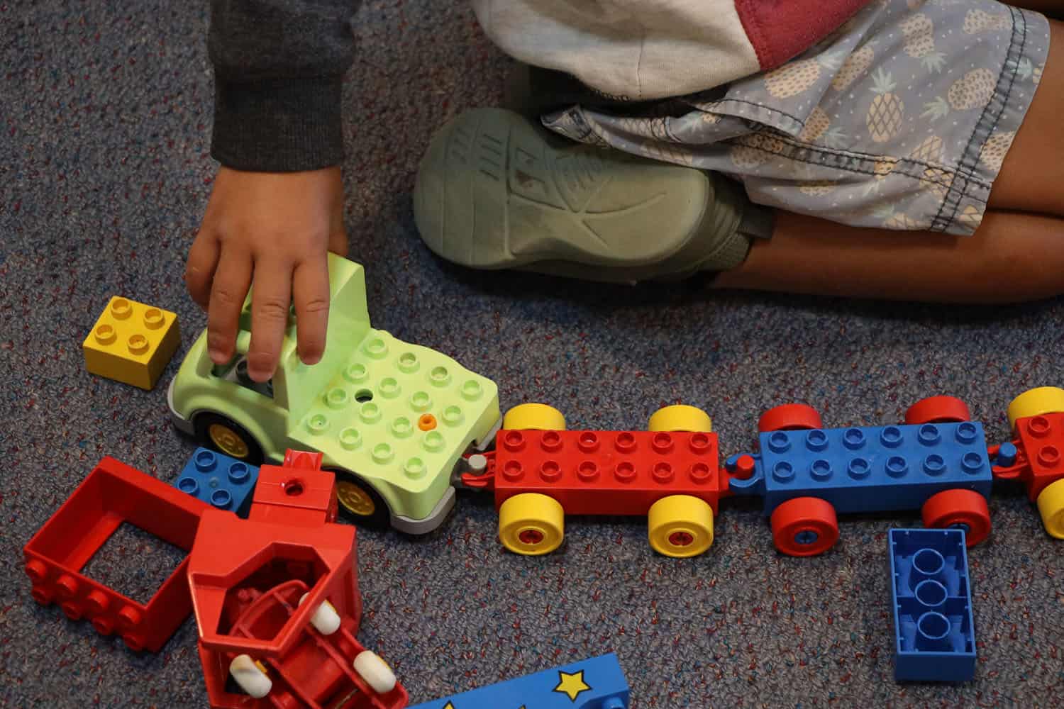 Child's hand is pushing toy car with trailers on floor of early childhood centre.