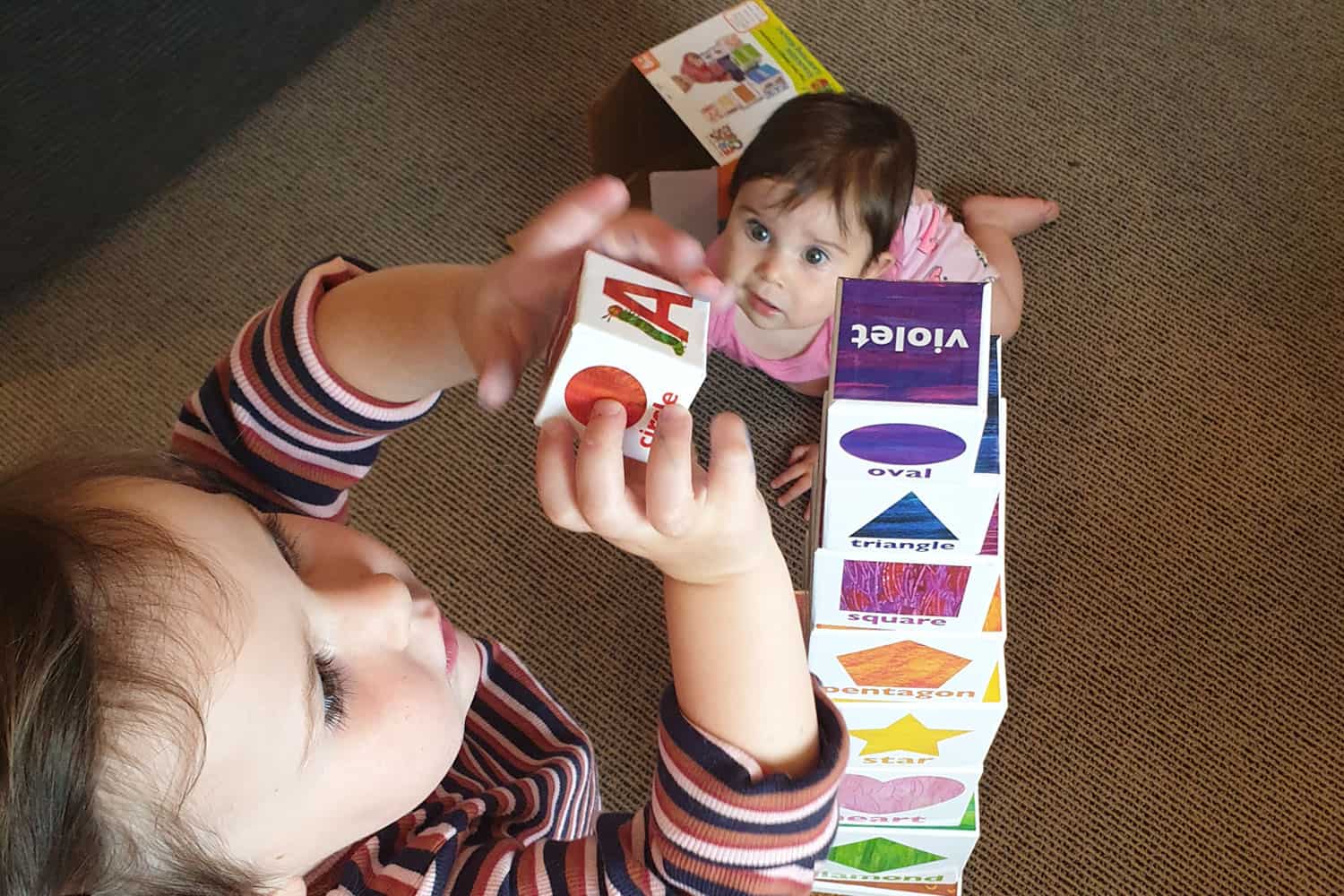 Young toddler with infant looking on, builds a tower of square blocks as tall as herself.