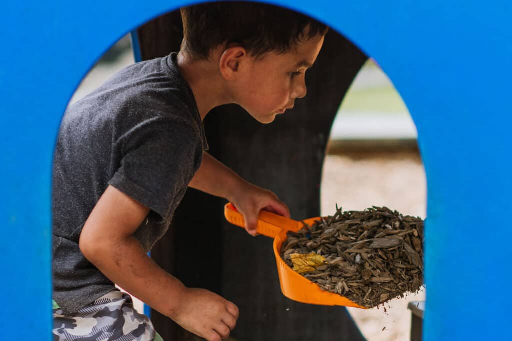 Boy busy in the preschool playground shovelling bark chips/ mulch.