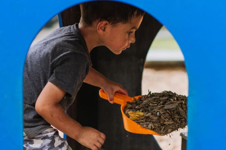 Boy busy in the preschool playground shovelling bark chips/ mulch.