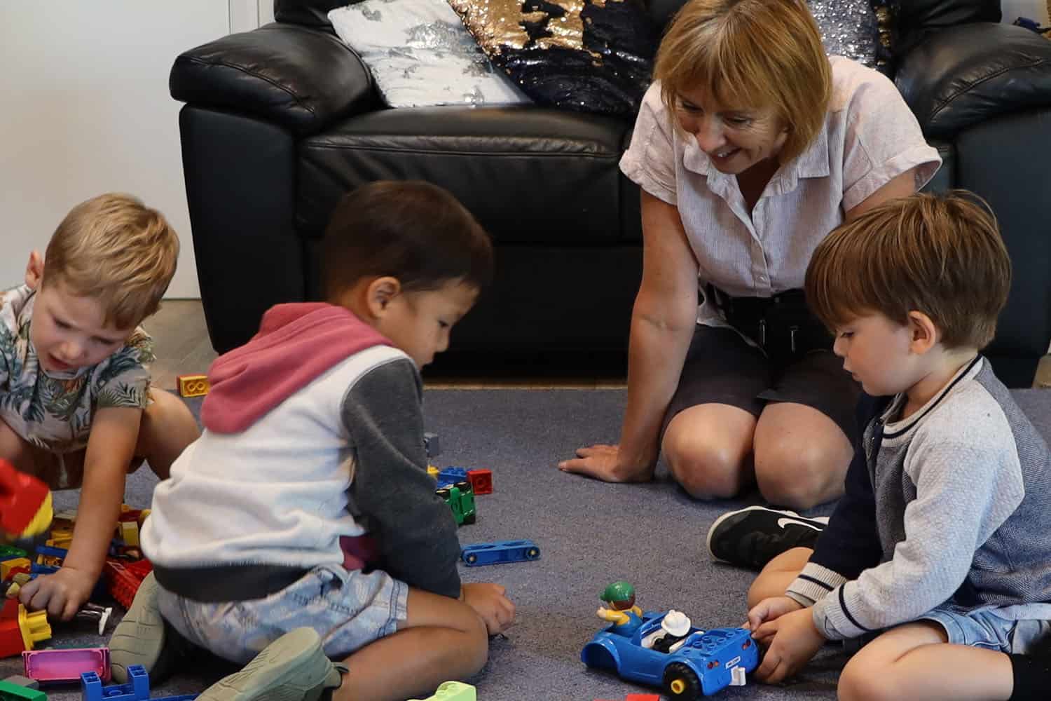 Teacher and three boys are playing with Duplo block toys on mat at early childhood centre.