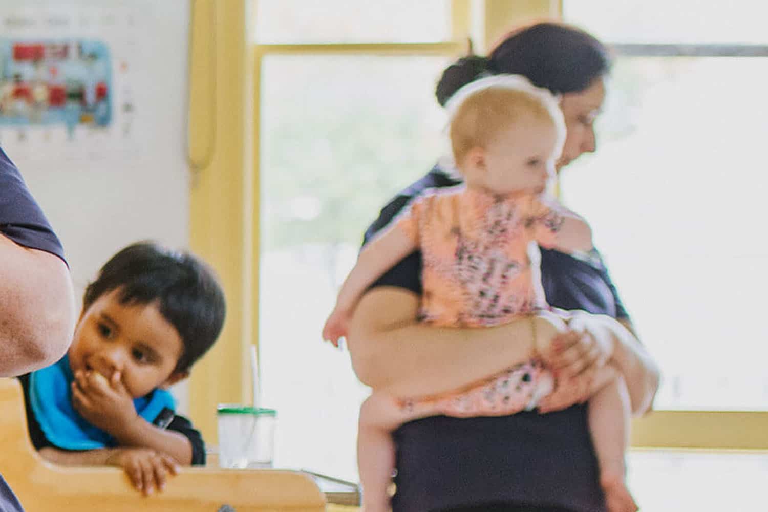 Toddler in a highchair eating and teacher holding an infant in lunch-room.