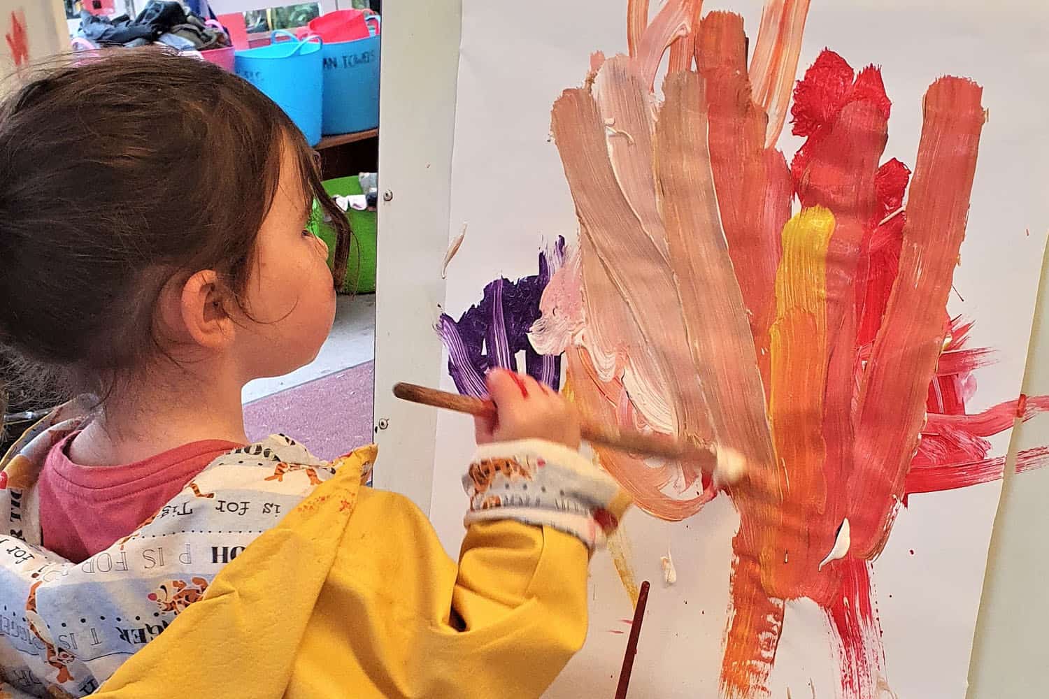 child with paint brush paints brightly coloured picture at kindergarten